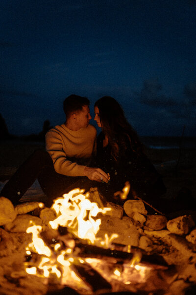 B & T staring into each others eyes about to kiss while in front of a campfire on the beach at night