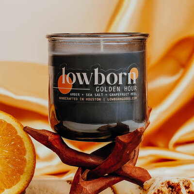 The moon butter candle, sitting on a pedestal covered in silky lilac fabric, the same fabric that is seen in the background.. The candle is surrounded by silver and gold abstract sculptures with purple light reflecting off of them. The image evokes smooth skin and a spacey spa day.