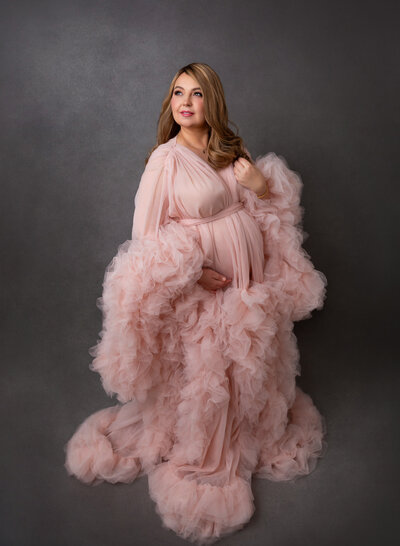Expectant Mom in pink organza and tulle down is standing with one arm resting under her bump and the other touching her shoulder. Mom is looking over her shoulder and smiling towards the light. Captured in Brooklyn, NY Maternity Photography Studio.