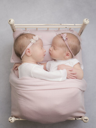 Twin new baby girls sleeping peacefully, snuggled together on a white metal bed. Wrapped in a pink blanket with bows, against a soft gray background. Captured in Matthews, NC for Charlotte Newborn Photographer Alicia Insley Smith