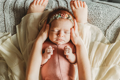 A peaceful newborn baby dressed in a pink onesie, with a floral headband, lies with its tiny hands next to the cheeks while peacefully sleeping on a soft, knit blanket during their in home newborn session in Harrisburg
