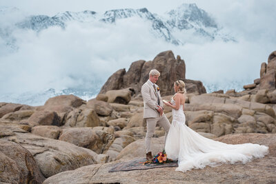 A bride in a long, full "poofy" wedding dress faces away from the luxury elopement photographer towards her partners as she reads vows during their California luxury elopement wedding.  He's wearing a khaki colored suit and a bright bouquet is visible on the rug they are standing on that's placed on a rock.  behind them rows of rocks with rounded edges are visible, and behind that is a low cloud slightly obscuring huge snow capped craggy mountains filling the back of the frame. 