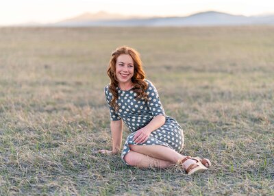 Senior session with a red-haired senior in a polka dot dress sitting in a field, warm evening light highlighting her natural beauty.