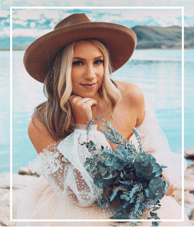 Woman sitting in a bridal dress by the lake, wearing a brown hat and holding a bouquet.