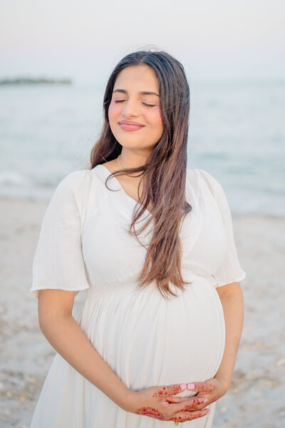 A soon-to-be mom stands on the beach in a white flowing gown, her eyes closed as she soaks up the warm sun, embracing the peaceful moment before welcoming her baby into the world.
