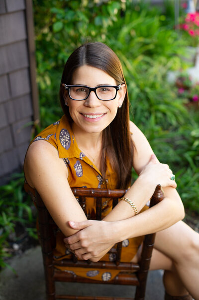 Creative headshot photo of an artist with mustard dress and glasses, sitting backwards in a chair with greenery in the background.
