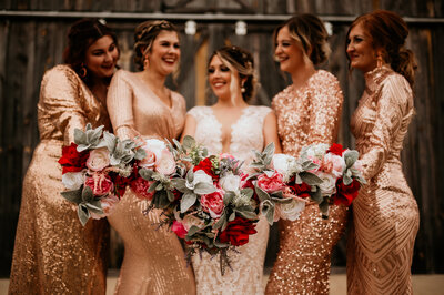bride in the middle of her bridesmaids who are in sequence gold gowns with cream and red floral bouquets captured by little rock ar wedding photographers