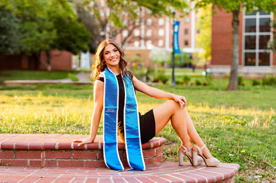 Senior girl in a black dress with her graduation stole on. She's sitting on the brick stairs outside the library