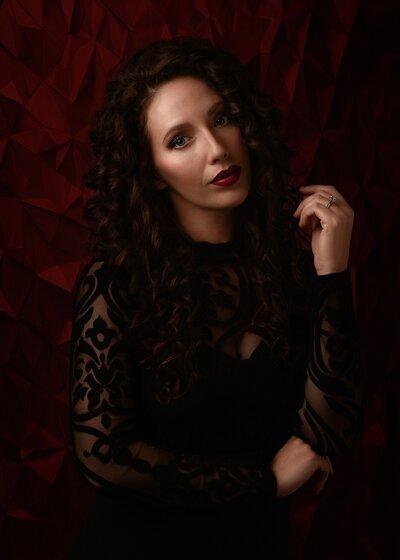 Amber Damour artist in black jumper with brown hair blue eyes and red lipstick on grey background