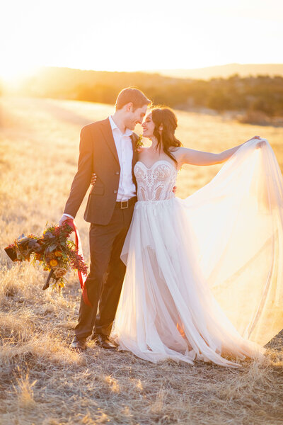 a bride and groom stand in an open field as the holds up the dress and the sun comes through