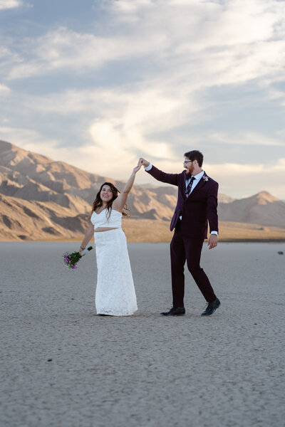 A man spins a woman on a dry lake bed. They are both smiling, she holds a bouquet of flowers