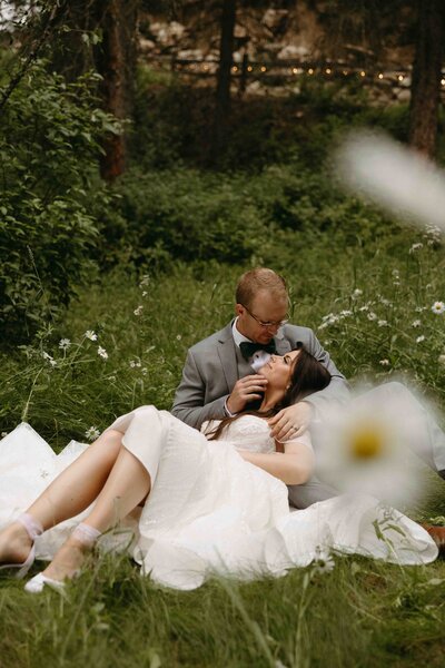 Bride and groom laying in the grass, with the bride in the groom’s lap looking up at him, surrounded by daisies.