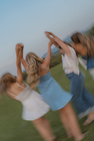 Four senior girls playfully hold their hands up in the air while holding hands with a slow shutter blur. There are mountain in the background and they are in a grassy field in Northeast, TN. 