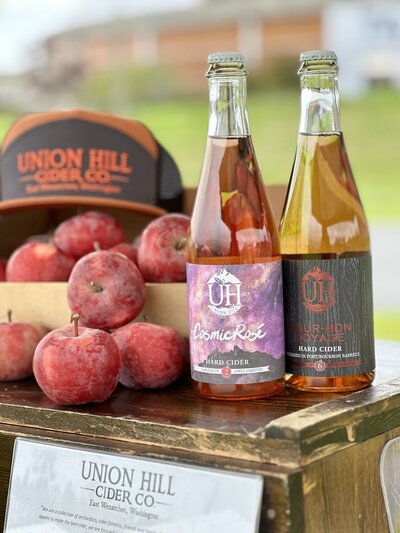 Two bottles of hard cider sitting on table next to some red apples and a Union Hill Cider Co trucker hat.