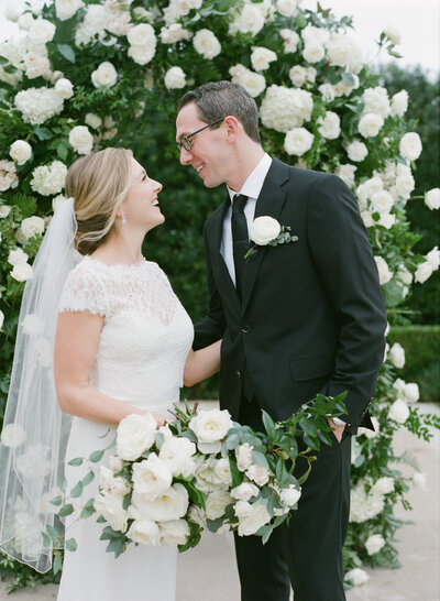 Bride and groom smiling at each other standing in front of a white and green floral arch