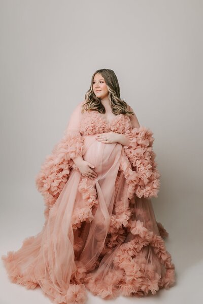 portrait of a woman taken in portland photography studio. She is wearing a pink dress and smiling over her shoulder. her hands are on her pregnant belly.