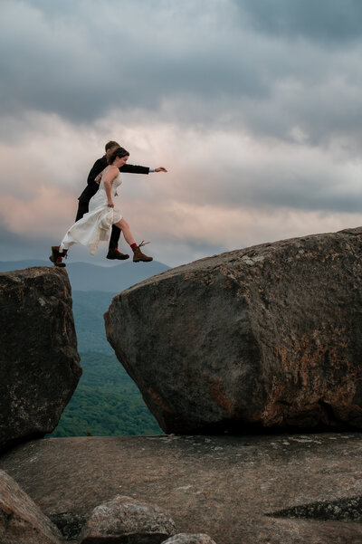 Couple jumps from one rock to another on a mountain summit at sunset in wedding attire.