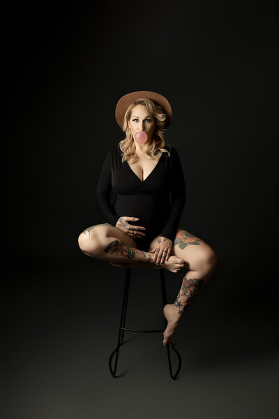 Expectant London,  Ontario mom at maternity photo session. Mom is sitting on a stool wearing a black, long sleeve bodysuit, her legs bare exposing tattoos. Her hair is loosely curled with a wide brimmed felt hat. She is blowing a big bubble with pink bubblegum. She is against a black backdrop.