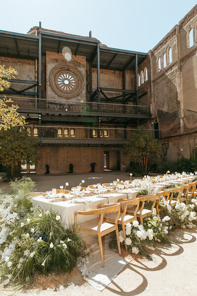 Newly weds stand together at the alter with their faces touching. They are surrounded by family with a flower bed and mountain scene in the background
