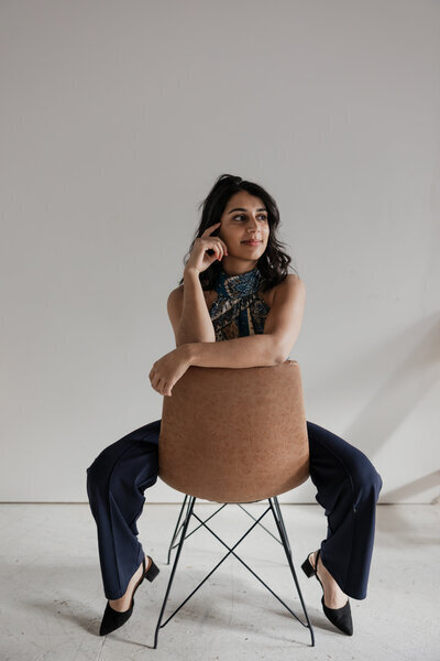 A person with long dark hair sits on a brown chair backward, resting their arms on the chair's back. They're dressed in dark pants and black heels, with a patterned top and a scarf adorning their neck—a classic style captured by a New England lifestyle photographer against a plain light-colored wall.