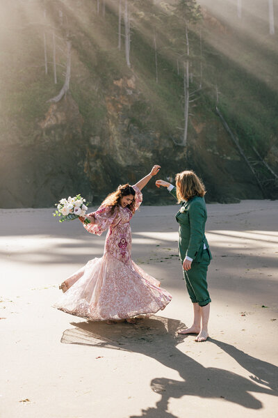 A groom in a green suit twirls his bride wearing a pink rue de Seine gown at their Oregon Coast elopement in Neskowin.