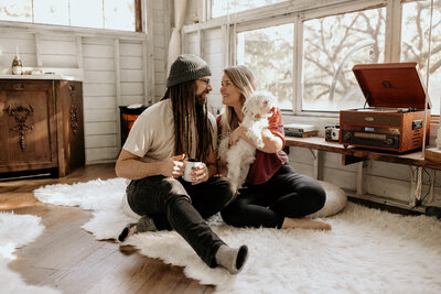 Man with dreads, beanie, and glasses smiles big at his wife while they both sit down on the floor. Wife is holding a little white fluffy dog with a beaming smile as she looks at her husband