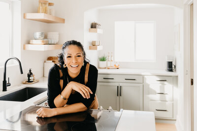Woman in a black sweater and leather apron leans on her kitchen counter
