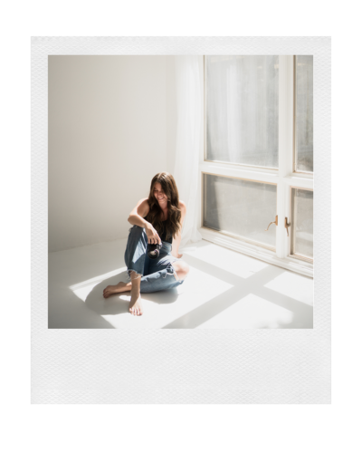 polaroid photo of a woman sitting on the floor in front of a big floor to ceiling window while holding a canon camera and looking down