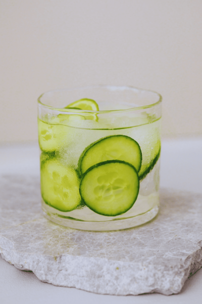 A glass of cucumber water with slices of cucumber and ice cubes. The glass is resting on a light-colored stone surface.