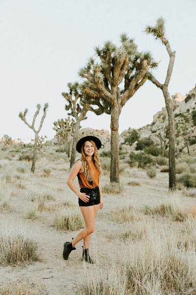 Laura smiling and standing near Joshua Trees, holding a camera looking happy.