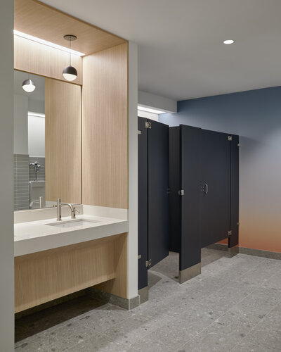 A washroom sink area featuring a bright white countertop,  sleek silver sink and soap dispenser, warm wood accents,  a painted wall gradient, and dark coloured stalls