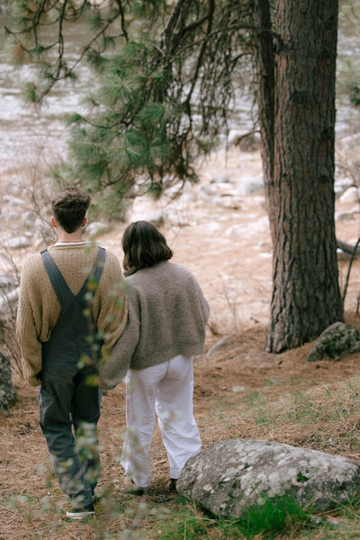 Couple walking hand in hand through a pine forest along the river during a lifestyle photography session in Idaho
