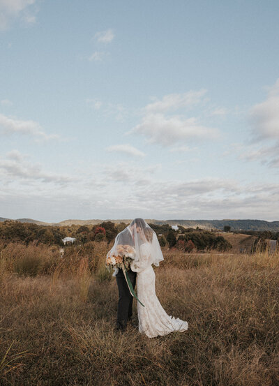 bride and groom in the middle of a fall field under the brides veil and embracing with a view of the mountains in Knoxville and a clear sky captured by Knoxville wedding photographers 