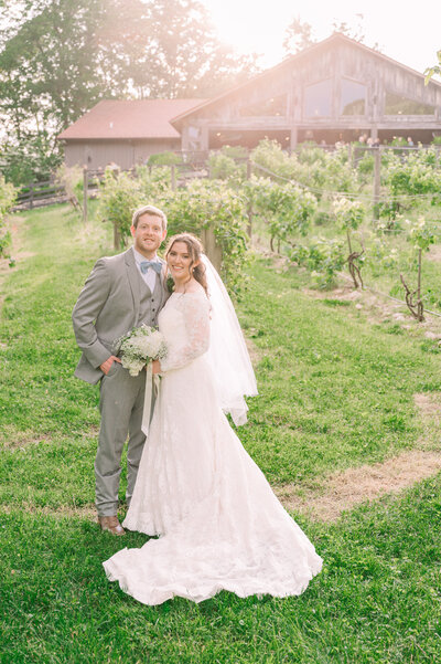 A smiling bride and groom embracing at sunset in June at The Vineyard at High Holly holding a white wedding bouquet by JoLynn Photography, a destination wedding photographer