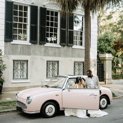 bride and groom posing with a pink car on their wedding day.