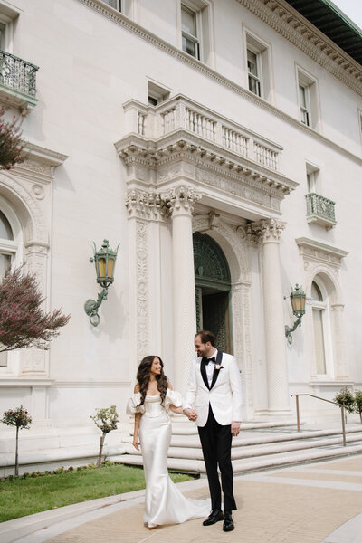 Bride is wearing a white couture wedding dress and groom is in a white tux and they are holding hands and walking in front of Flood Mansion in San Francisco California