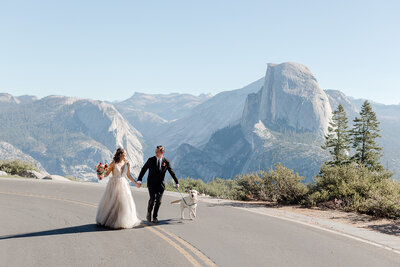 A bride and groom run down an iconic Yosemite road with their dog, all in view of Half Dome. 