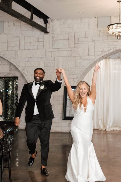 bride and groom enter their wedding reception holding hands, with the bride raising her arms in celebration