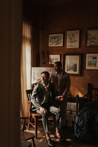 Two partners share an intimate moment in a warmly lit room during their downtown Tucson, Arizona wedding. One partner is seated, while the other stands beside them, resting a hand on their shoulder. The room is adorned with framed artwork and vintage decor.