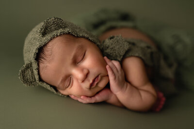 Dressed in a charming bear bonnet and surrounded by earthy green tones, this newborn rests peacefully in a pose that captures gentle serenity.