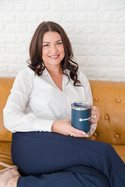 The professional in body contouring services Dallas wears an elegant outfit and sits on a brown office couch.