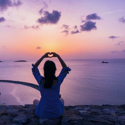 Female sitting in front of a sunset over the water with her hands in the air in the shape of a heart