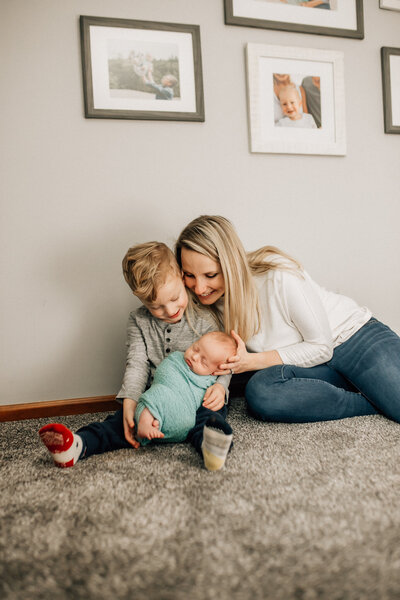 Newborn photography photoshoot session with mom, young boy, and baby sitting on the floor smiling.