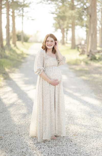 Pregnant woman in a neutral floral dress walking along a wooded path at sunset during a maternity session with a Kansas City maternity photographer.