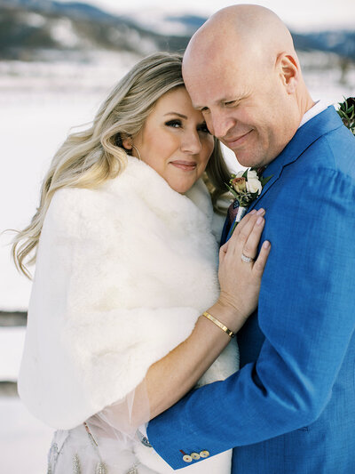 Close up of a bride and groom embracing. The bride has long blond hair, and is wearing a white fur shall. The groom is wearing a blue suit.