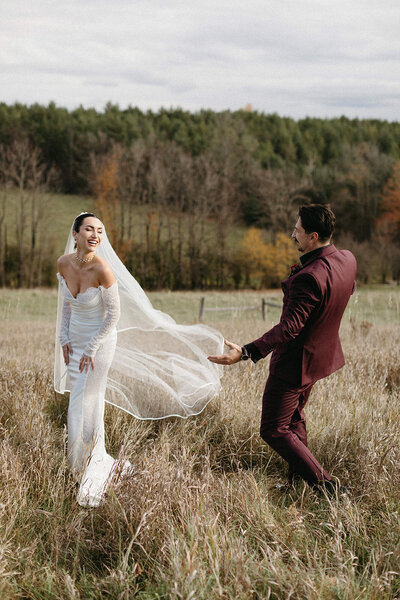 A bride in a white dress laughs as she walks through a grassy field, captured beautifully by Toronto wedding photographer, Jennifer van Son, at Farmhill weddings in Peterborough. Her groom in a burgundy suit follows her, framed by trees and the cloudy sky, showcasing the charm of wedding photography in Toronto.