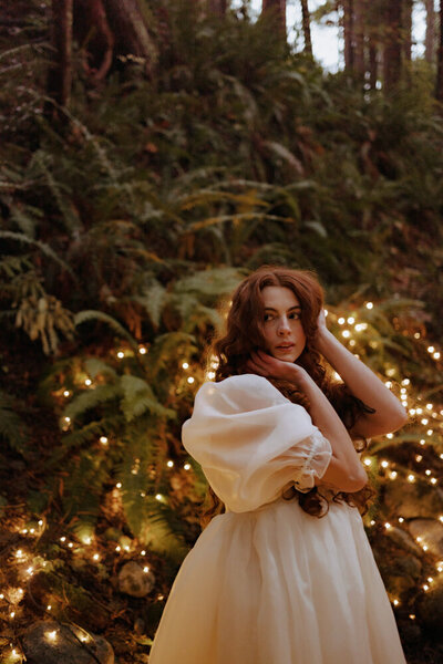 Red headed woman in a white dress stares off into the distance while posing naturally in front of pine trees covered in white fairy lights