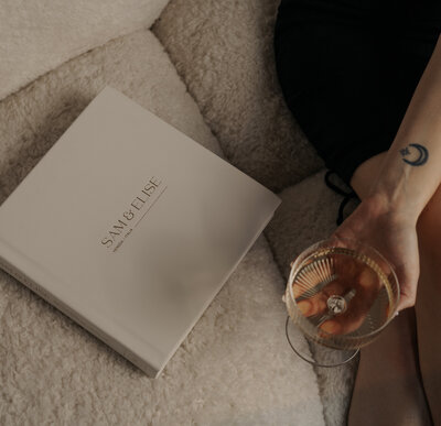 Woman with black dress holding mix drink sitting on couch  next to large white wedding album with the name sam & elise debossed on the cover- Romero Album Design