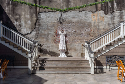 A white lace wedding dress hangs on display between two matching staircases against a weathered brick wall, with a black chandelier and greenery garland above. Sunlight casts shadows on the ground.
