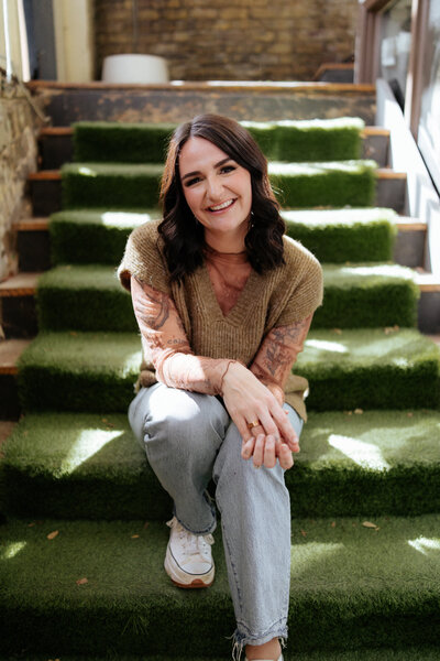 a brunette woman sitting on green stairs smiling while wearing light colored jeans and a tan sweater.